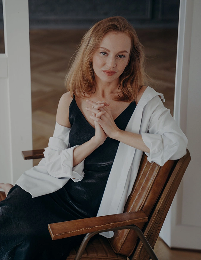 Woman with red hair in white blouse and black vest sitting on wooden chair with hands clasped