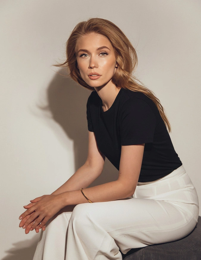 Woman in black t-shirt and white pants sitting on stool against neutral background in minimalist portrait
