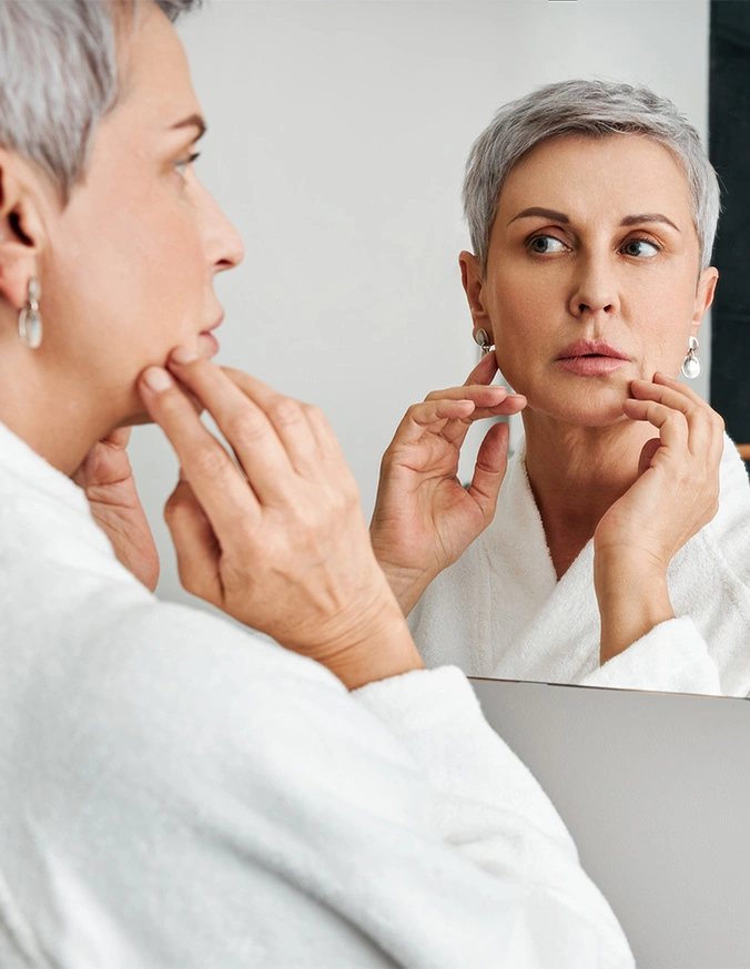 Woman examining facial skin in mirror while wearing white robe, checking for signs of aging