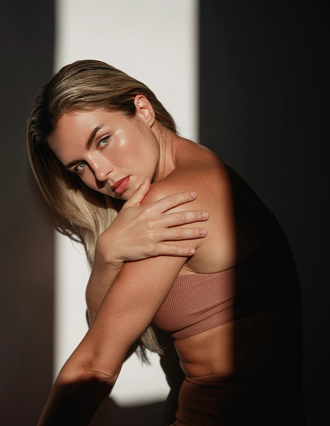 Blonde woman in beige tube top posing against white wall with dramatic shadow lighting