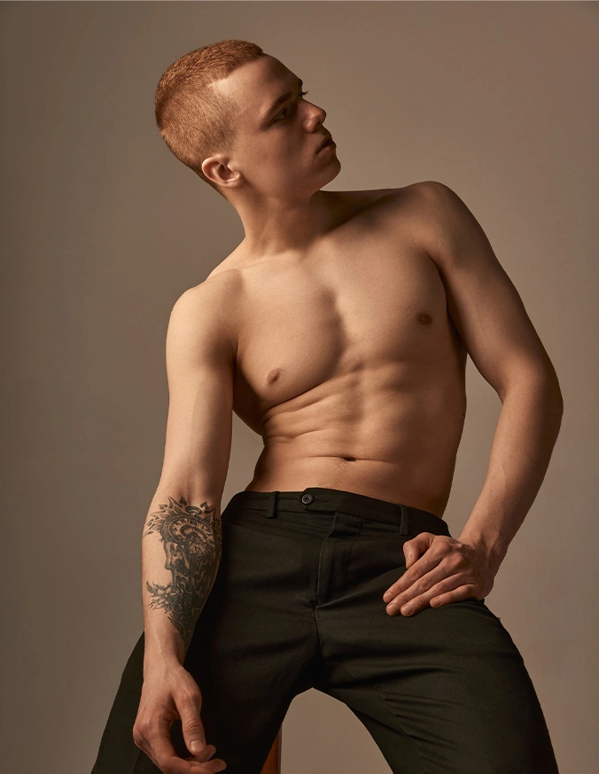Shirtless male model with tattoo posing in black pants against neutral background in fashion photography