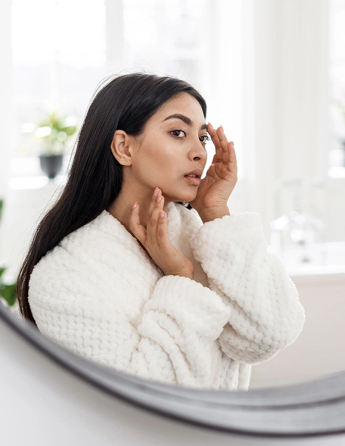 Woman in white bathrobe examining skin in mirror during skincare routine in bright bathroom