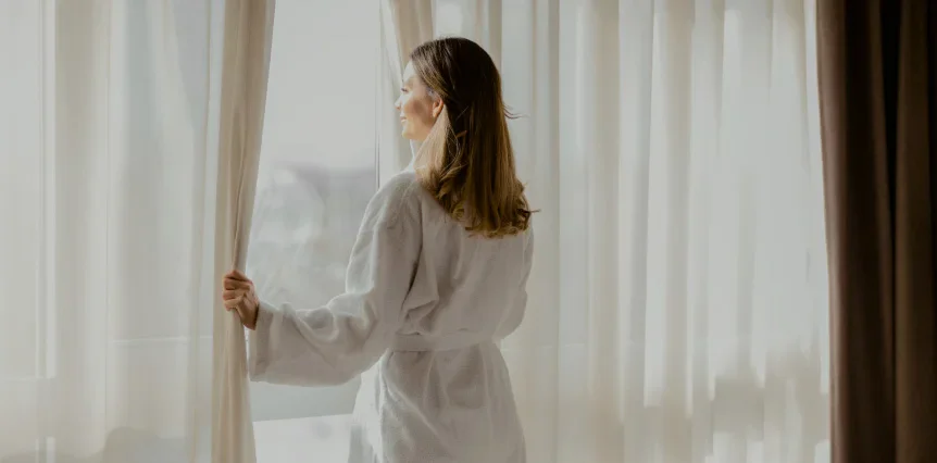 Woman in white robe opening curtains by sunny window in peaceful morning routine at home