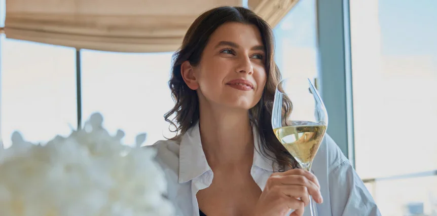 Woman enjoying white wine by window with city view and white flowers in foreground